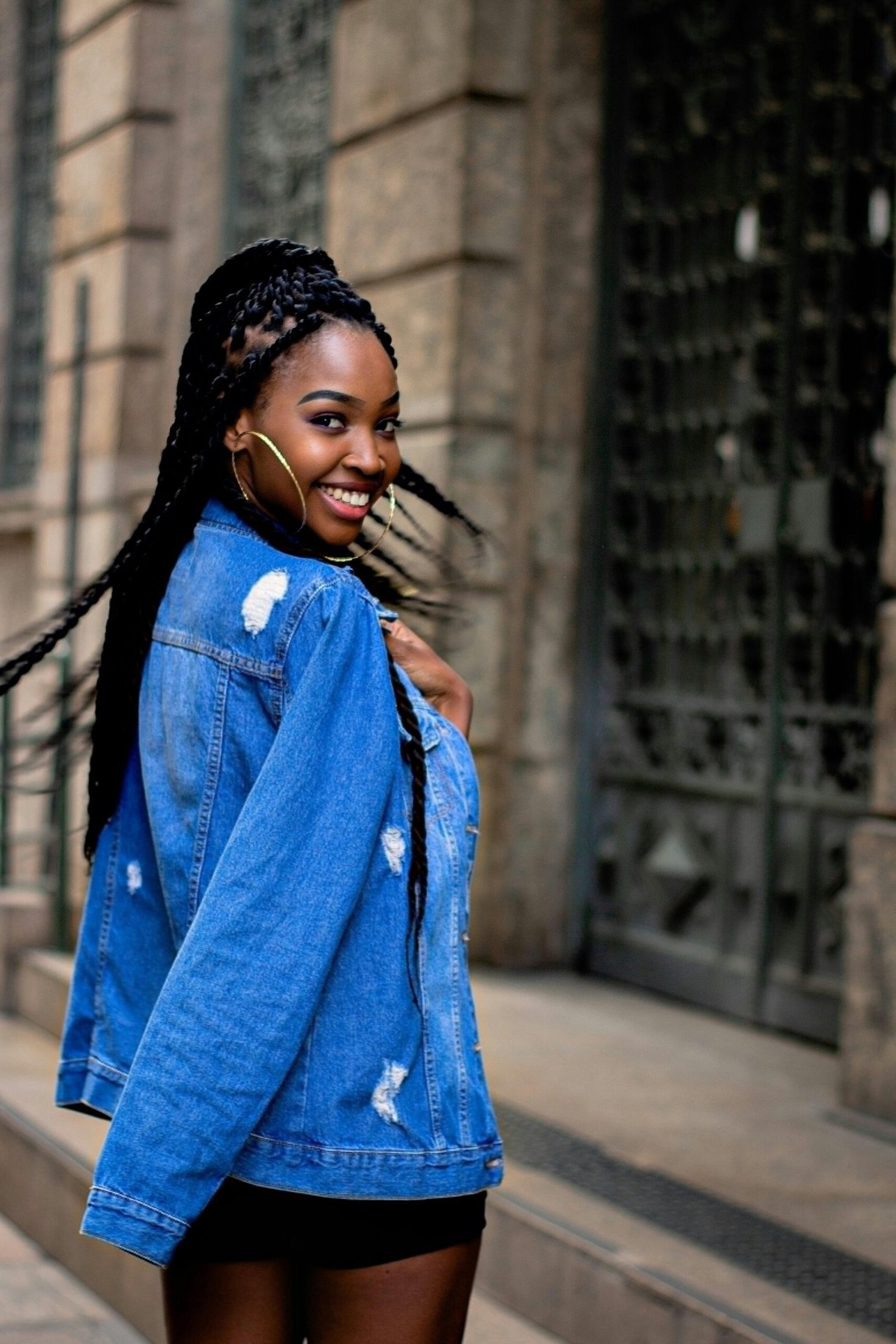 Stylish woman with braided hair smiling outdoors in denim jacket.
