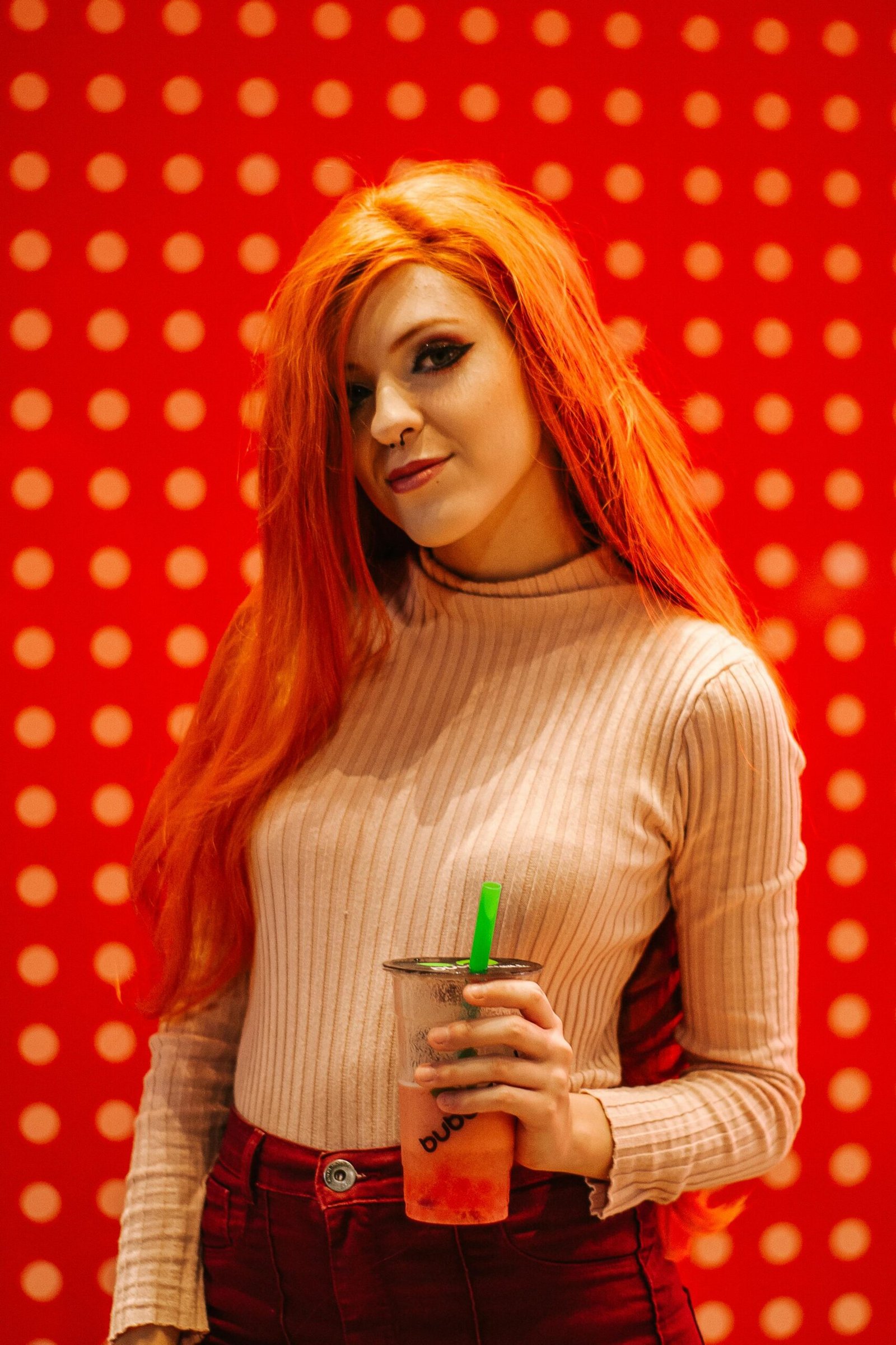 Woman with red hair holding bubble tea against vibrant polka dot background.